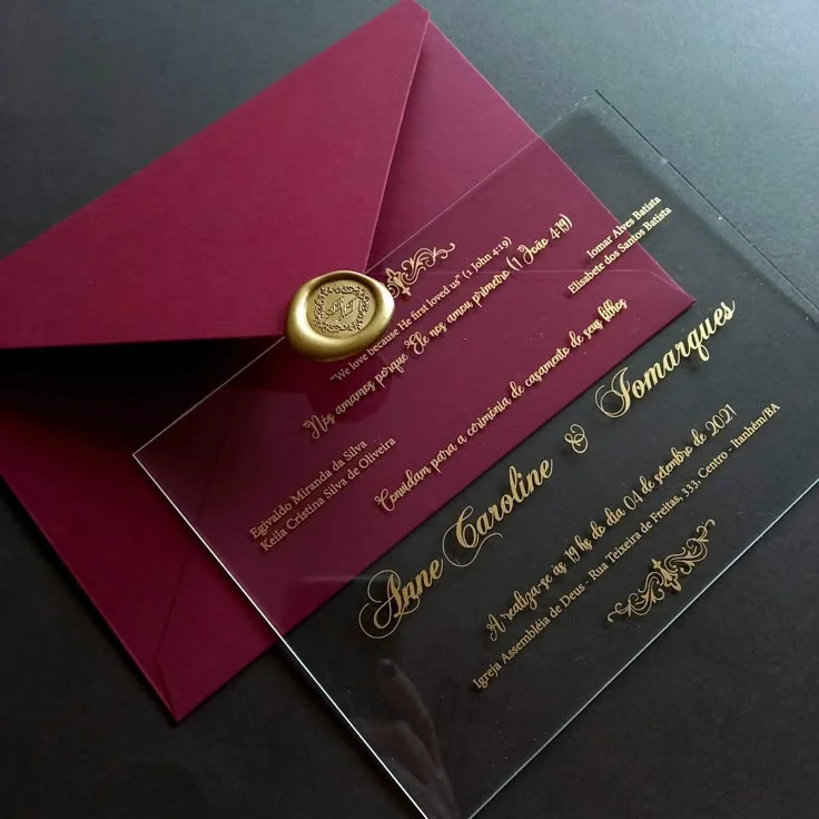 Transparent wedding invitation with gold lettering beside a burgundy envelope
